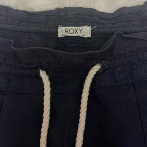Mörkblå linnebyxor från Roxy - Snygga mörkblå linnebyxor från Roxy med bred resår i midjan och grov vit snörning framtill. Byxorna har en lös passform och raka ben, perfekta för chill dagar. Materialet är linne som ger en skön och luftig känsla