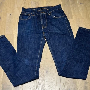 Mörkblå slim jeans från Nudie Jeans - Säljer ett par mörkblå jeans från Nudie Jeans med klassisk femficksdesign och kontrastsömmar. Byxorna har raka ben och snygg broderad detalj på bakfickorna. Tillverkade i slitstark bomullsdenim, perfekta för en clean och stilren look. I storleken W27 L32