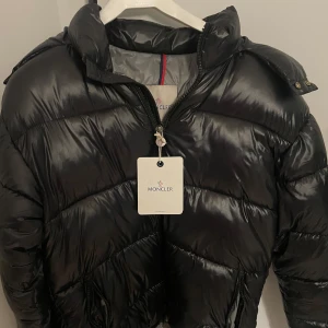  Moncler maya - Säljer en svart pufferjacka från Moncler med glansig finish och klassisk logga på ärmen. Jackan har dragkedja framtill, hög krage och flera fickor. Insidan har ett unikt serietryck. Perfekt för dig som vill ha en snygg och varm jacka med streetkänsla.