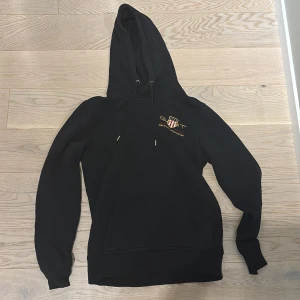 Svart hoodie från GANT med tryck - Svart hoodie från GANT med broderad logga och text på bröstet. Tröjan har huva med snörning, magficka och långa ärmar med ribbade muddar. Perfekt för chill dagar och snygg streetstyle. Materialet känns mjukt och är troligen bomull eller polyester.