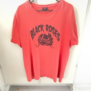 Röd Black Roses t-shirt Adam XL - Cool röd t-shirt från Adam i storlek XL med trycket 'Black Roses' och en svart ros med döskalle på bröstet. T-shirten har rund hals och korta ärmar, perfekt för dig som gillar edgy streetstyle. Materialet är mjuk bomull som känns skönt mot huden.