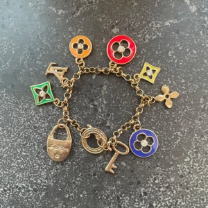 Vintage armband  - Superfint armband med flera färgglada berlocker i guldmetall. Charmsen har lås, nyckel och blommor i rött, grönt, blått, gult och orange. Kedjan är i guldfärgad metall och detaljerna har glittrande stenar i mitten. Perfekt statement piece för din look!