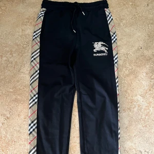 Svarta Burberry mjukisbyxor med rutig kant - Svarta mjukisbyxor från Burberry med klassisk rutig kant längs sidorna i beige, svart, vit och röd. Byxorna har snörning i midjan och broderad Burberry-logga framtill. Skön passform och stilren look som sticker ut med de rutiga detaljerna.