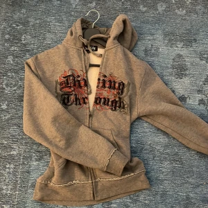 Grå hoodie från Identity med tryck - Säljer en grå hoodie från Identity med coolt tryck i svart och rött på framsidan. Tröjan har huva med snörning, dragkedja och långa ärmar. Snygga råa sömmar och mjukt material som känns skönt mot huden. Perfekt för dig som gillar streetstyle. Finns små fettfläckar som säkert går bort lätt