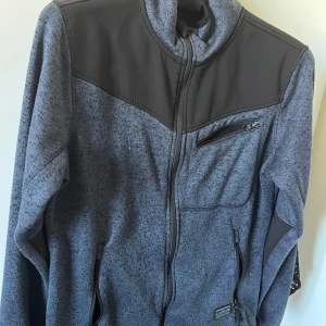 Grå fleecejacka - Snygg grå fleecejacka från Bluewear med svarta detaljer på axlar och armbågar. Den har ett litet hål längst ner på ärmen som nog är lätt att sy igen. Jackan har hel dragkedja, tre praktiska fickor med dragkedja och en hög krage. Perfekt för dig som gillar en sportig och chill stil.