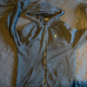 Svart hoodie från Lyle & Scott - Svart hoodie från Lyle & Scott med gul broderad logga på bröstet. Tröjan har dragkedja framtill, huva och två fickor. Tillverkad i mjuk bomull och har långa ärmar. Perfekt för chill dagar och streetwear-stil.