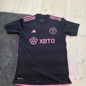 Inter Miami CF svart fotbollströja - Svart fotbollströja från Inter Miami CF med rosa detaljer och klubbmärke på bröstet. Adidas-logga och sponsortryck i rosa framtill, samt tre rosa ränder på axlarna. Tillverkad i lätt och ventilerande polyester med subtila vågmönster i tyget.
