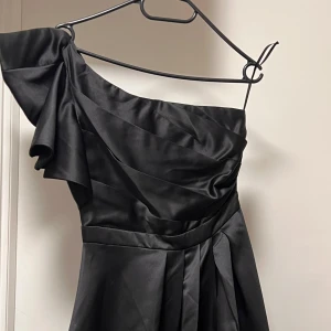 Svart långklänning med oneshoulder - Supersnygg svart långklänning med oneshoulder och volangdetalj på axeln. Klänningen har ett elegant, draperat liv och ett brett midjeband som ger en markerad siluett. Kjolen är vid och faller snyggt i mjukt, glansigt tyg som ger en lyxig känsla.