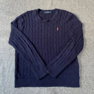 Mörkblå kabelstickad tröja från Polo Ralph Lauren - Säljer en stilren mörkblå kabelstickad tröja från Polo Ralph Lauren med det klassiska logotypbroderiet i orange på bröstet. Tröjan har långa ärmar och ribbade kanter vid ärmslut och nederkant. Perfekt för kyligare dagar! Priset är diskuterbart.