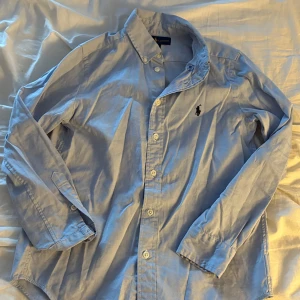 Blå skjorta från Ralph Lauren - Snygg ljusblå skjorta från Ralph Lauren med klassisk krage och knappar framtill. Broderad svart logga på bröstet. Långärmad och tillverkad i mjuk bomull. Perfekt för dig som gillar stilrena och tidlösa plagg.