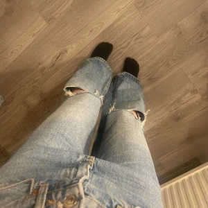 Blå wide jeans  - Säljer ett par ljusblå jeans med wide fit från gina tricot. De har hög midja och slitna detaljer med stora hål på knäna för en cool, avslappnad vibe. Klassisk femficksmodell och raka ben. Perfekta för dig som gillar streetstyle och oversized look.