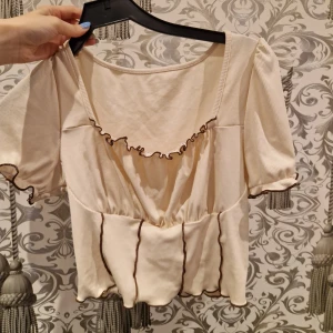 Beige blus med volang och bruna sömmar - Supersöt beige blus med korta puffärmar och bruna kontrastsömmar. Blusen har volangdetaljer vid halsringningen och längs kanterna, samt en lätt rynkad midja som ger en chill och feminin vibe. Perfekt för dig som gillar romantisk och trendig stil.