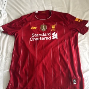 Liverpool FC röd matchtröja 2019 - Liverpool FCs officiella röd matchtröja från 2019 med vita tunna ränder, broderat klubbmärke och FIFA World Champions 2019-emblem. Kortärmad med vit kant vid kragen och ärmslut, tillverkad i lätt och ventilerande polyester. Snyggt tryck från Standard Chartered på bröstet.