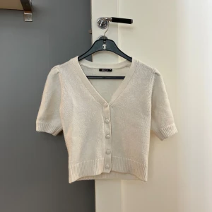 Vit stickad kofta från Gina Tricot - Supersöt vit stickad kofta från Gina Tricot i storlek XS. Kort modell med v-ringning, ribbade muddar, fem matchande knappar framtill och puffiga korta ärmar.