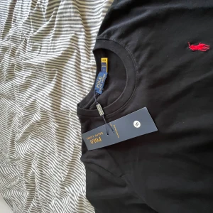 Svart t-shirt från Polo Ralph Lauren - Svart t-shirt från Polo Ralph Lauren med klassisk röd logobroderi på bröstet. Rund hals och korta ärmar, tillverkad i mjuk bomull. Enkel och stilren design som funkar till jeans eller shorts. Perfekt för dig som gillar clean och tidlös look.