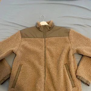 Beige teddyjacka från Lindex - Mysig teddyjacka från Lindex i beige med kontrasterande paneler i brunt upptill och vid fickorna. Jackan har hög krage, hel dragkedja och två sidofickor. Perfekt för kalla dagar med sin fluffiga och sköna känsla. Passformen är normal och jackan är långärmad.