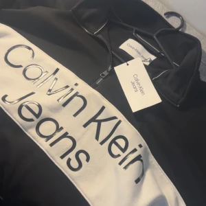 Svart half zip sweatshirt Calvin Klein Jeans - Skicka dm för video/prisförslag. Svart sweatshirt från Calvin Klein Jeans med vit panel och stor logga framtill. Tröjan har half zip, ståkrage och långa ärmar med elastiska muddar. Snygg och enkel design som passar till jeans eller joggers. Materialet känns som mjuk bomull med inslag av polyester.