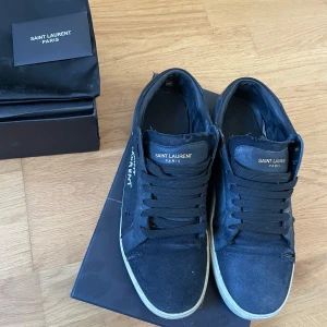 Svarta Saint Laurent sneakers - Svarta sneakers från Saint Laurent med vit logga på sidan och svart logga på plösen. Skorna har snörning, rund tå och platt vit sula. Materialet är canvas och skinn, vilket ger en cool och urban vibe. Perfekt inför hösten. Defekter syns på bilderna. Priset ej hugget i sten! Allt på bilderna ingår.