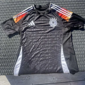  Tysklands fotbollströja  - Säljer en svart Tysklands fotbollströja från Adidas med vita och färgglada ränder på axlarna. Tröjan har korta ärmar, rund hals och Tysklands landslagsemblem med fyra stjärnor på bröstet.