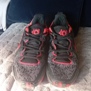 Nike KD 15 EP 'Bred' - Säljer ett par Nike KD sneakers i svart och rött med coolt mönstrad sula och tydlig KD-logga på plösen. Skorna har snörning, mesh-överdel och en låg siluett. Swoosh-loggan på sidan och detaljer i rött ger dem en sportig vibe. Perfekta för dig som gillar basket eller streetstyle.