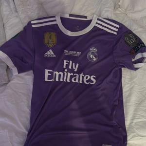 Real Madrid Marcelo 12 lila matchtröja - Säljer en officiell Real Madrid Champions League-tröja från finalen i Cardiff 2017, med Marcelo och nummer 12 på ryggen. Tröjan är lila med vita detaljer, Adidas-logga, Fly Emirates-tryck och Champions League-patchar på ärmarna. Materialet är lätt polyester med climacool-teknik. Köpt för 500kr säljs för 250