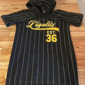 Svart randig hoodie Légalité - Svart hoodie med vita ränder och gul text 'Légalité EST. 36' på bröstet. Tröjan har korta ärmar, huva och en stor magficka framtill. Perfekt för dig som gillar streetwear och vill sticka ut med coola detaljer.