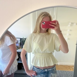 Ljusgul blus med volangmidja - Säljer en ljusgul blus med korta ärmar och rund hals. Toppen har en snygg smockad midja som ger en volangig och feminin siluett. Perfekt till jeans eller kjol för en fräsch look. Materialet känns mjukt och lätt, troligen bomull.