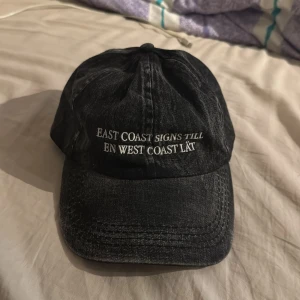 Hov1 keps - Snygg svart keps i jeansmaterial med vit broderad text på framsidan: 'EAST COAST SIGNS TILL EN WEST COAST LÅT'. Från ett gammalt hov1 släpp 
