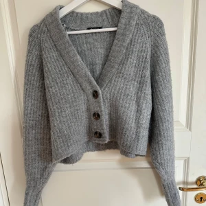 Grå stickad kofta med knappar - Mysig grå stickad kofta från Gina Tricot med ribbad struktur och fyra stora knappar framtill. Koftan har en djup v-ringning, lång ärm och är lite croppad i modellen. Perfekt för lager-på-lager och chill dagar.