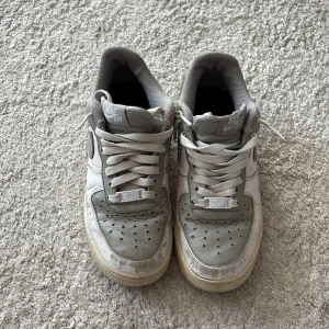 Nike Air Force 1 grå sneakers - Nike Air Force 1 sneakers i grått och vitt med klassisk låg siluett. Skorna har snörning, rund tå och platt sula. Ovandelen är i skinn med mesh vid kragen för extra komfort. Swoosh-logga på sidan och Nike Air-brodyr på hälen. Egen designad sko. Original sko köpt på Nikes egna hemsida  