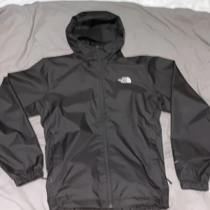Svart vindjacka från The North Face - Svart vindjacka från The North Face med huva och dragkedja framtill. Jackan har elastiska muddar vid ärmslut och nederkant, samt logga på bröstet och ryggen. Tillverkad i lätt polyester som är perfekt för blåsiga dagar. Enkel och clean design som funkar till streetwear. Knappast använd!