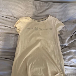 Beige t-shirt från Sail Racing - Beige t-shirt från Sail Racing med rund hals och korta ärmar. Framsidan har diskret Sail Racing-logga i grått och baksidan har en liten logodetalj vid nacken. T-shirten är gjord i mjuk bomull och har en clean, enkel look som funkar till jeans eller shorts. Knappt använt, tröjan är även äkta.