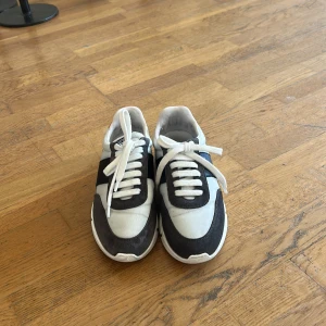 Axel Arigato sneakers i grå och vit - Snygga sneakers från Axel Arigato i storlek 40. Skorna har en fräsch mix av grå mocka och vit textil, med coola detaljer i orange på hälen. Klassisk snörning och rund tå. Perfekta för dig som gillar stilrena och trendiga sneakers.