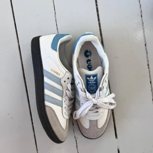 Adidas Samba sneakers vit/blå - Säljer ett par trendiga Adidas Samba sneakers i vitt med ljusblå ränder och detaljer. Skorna har svart platt sula, snörning och rund tå. Ovandelen är i skinn och mocka. Skorna är knappt använda och är i ett väldigt bra skick🩵🩵