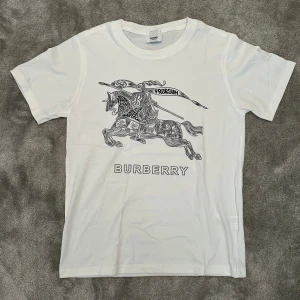 Vit Burberry t-shirt - ✨ Burberry T-shirt ✨ Snygg vit t-shirt från Burberry 🤍 med ett stort grafiskt tryck av en riddare på häst och texten “BURBERRY” framtill ⚔️. 	•	👕 Rund hals och klassisk passform 	•	🧵 Tillverkad i 100% bomull för mjuk och skön känsla 	•	🌟 Perfekt för dig som gillar stilrena designer med coola detaljer  En t-shirt som kombinerar ikonisk stil och moderna detaljer.