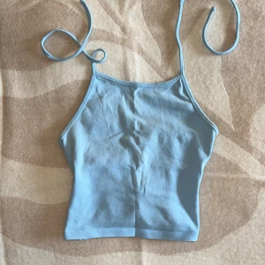 Ljusblå croppad linnetopp med knyt - Supersöt ljusblå croppad linnetopp med tunna axelband som knyts i nacken. Toppen har en enkel och stilren design, perfekt för att matcha med jeans eller kjol. Mjuk bomullskänsla och lätt tyg som passar när det är varmt ute.