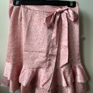 Rosa kjol med volanger - Supersöt rosa omlottkjol med vitt prickigt mönster och volanger längst ner. Kjolen har en knytning i midjan som ger en snygg detalj och sitter bekvämt. Perfekt för dig som gillar ljusa färger och lekfull stil.