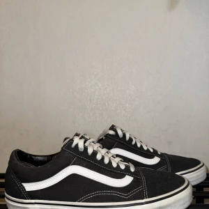 Svarta Vans Old Skool sneakers 43 - Klassiska Vans Old Skool sneakers i svart med vit sidorand och vita snören. Skorna har en låg profil, rund tå och platt sula. Ovandelen är i slitstark canvas och mocka, med ikonisk Vans-logga på sidan. Perfekta för dig som gillar streetstyle och skate. 