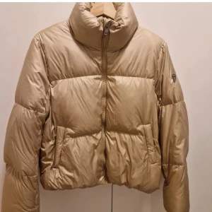 Beige pufferjacka Rockandblue 38 - Snygg beige pufferjacka från Rockandblue med hög krage och dragkedja framtill. Jackan har en cool badge på ärmen och rosa foder på insidan. Perfekt för kalla dagar och har en loose passform som ger en chill vibe. Den är använd jätte få gånger. 