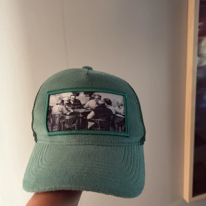 Grön keps från Emilio Pendenza - Snygg grön keps från Emilio Pendenza med mesh på sidorna och baksidan för extra ventilation. Framtill finns en svartvit patch med motiv av personer som sitter vid ett bord. Justerbar baktill och cool streetstil.