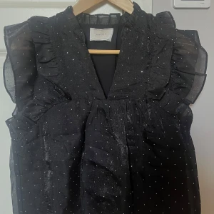 Svart glittrig blus från Neo Noir - Svart blus från Neo Noir med glittrigt tyg och små diamant detaljer. Toppen har volangdetaljer vid axlarna. Super söt men passade tyvärr inte mig och den är helt oanvänd har bara provats.
