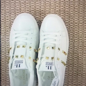 Valentino Rockstud vita sneakers - Säljer ett par vita Valentino Rockstud sneakers med coola guldfärgade nitar på sidorna och ovansidan. Skorna har snörning, platt sula och rund tå. Märket syns tydligt på både plösen och sulan. Perfekta för dig som gillar lyxig streetstyle.