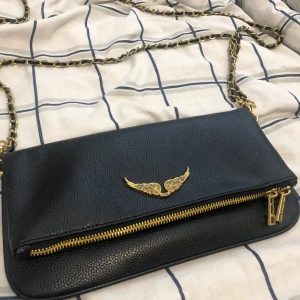 Svart axelväska från Zadig & Voltaire - Snygg svart axelväska i skinn från Zadig & Voltaire med guldfärgade detaljer, kedja och dragkedja. Framsidan har ett coolt emblem med vingar i guld och glittriga stenar. Perfekt storlek för mobilen och det viktigaste. Kedjan har inslag av svart läder.