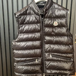 Moncler väst - Snygg svart dunväst från Moncler med glansig finish och klassisk logga på bröstet. Västen har ståkrage, två snedställda fickor med dragkedja och quiltad design. Perfekt för lager-på-lager och har en modern, clean look. Västen är sparsamt använd och självklart äkta! Storlek TG 3 vilket motsvarar M-S