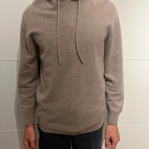 Beige hoodie i kashmir med huva - Mjuk och lyxig beige hoodie i kashmir med huva och snörning. Tröjan har långärmade ribbade muddar och en klassisk passform som är både chill och stilren. Perfekt för dig som gillar en clean look och vill ha något extra skönt till hösten och vintern.   (Stor i storleken)