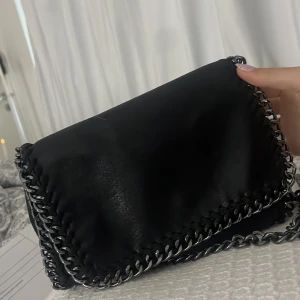 Svart axelväska med kedjedetaljer - Snygg svart axelväska med grova silverfärgade kedjor runt kanterna och som axelrem. Väskan har en stilren, rektangulär form och magnetlås. Materialet är glansigt skinnliknande och ger en cool vibe. Perfekt för dig som gillar edgy accessoarer.