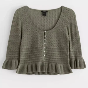 Olivgrön stickad kofta från Lindex - Säljer en olivgrön stickad kofta från Lindex med snyggt mönster och volangkant vid ärmslut och nederkant. Koftan har rund hals, knappar framtill och trekvartsärm. Perfekt för dig som gillar detaljer och mjuka material.