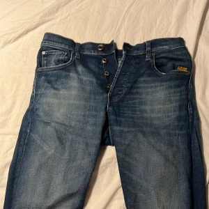 Blå jeans från G-Star RAW, W29 L32 - Snygga blå jeans från G-Star RAW med klassisk femficksdesign och knappgylf. Jeansen har en cool tvätt med ljusare partier och kontrastsömmar. Passar dig som gillar streetwear och vill ha ett par jeans med lite edge. Materialet är bomull med lite stretch för skön passform.