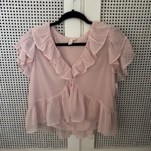 Ljusrosa blus med volanger från Nelly - Strl 36, aldrig använd. Supersöt ljusrosa blus från Nelly med volanger längs halsringningen, ärmarna och nederkanten. Blusen har korta ärmar och knappar framtill. Materialet är lätt och luftigt, perfekt för en romantisk look. Passar dig som gillar feminina detaljer och pastellfärger.