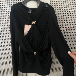 Svart party topp från SHEIN - Aldrig använd. Svart långärmad festtopp från SHEIN med snygga omlottdetaljer och två guldfärgade ringar framtill. Toppen har en cool twistad look och sitter tajt över kroppen. Perfekt för dig som gillar statement-plagg med lite edge.